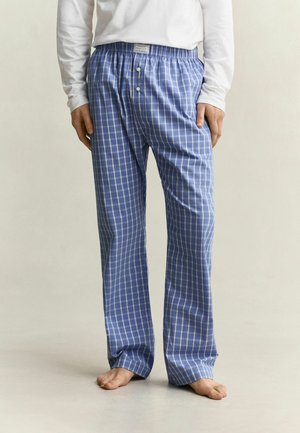 Persoon draagt blauw-wit geruite pyjamabroek met een wit langemouwenshirt en staat blootsvoets op een lichte vloer.