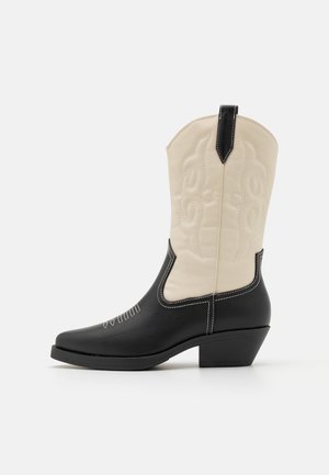 Kaksivärinen musta ja kerma cowboysaapas koristeompeleilla ja matalalla viistolla korolla, esitetty sivuprofiilissa valkoisella taustalla.