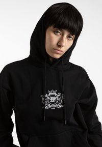 Felpa nera in cotone con tasca a marsupio frontale, caratterizzata da un design con stemma ricamato bianco e cordini sul cappuccio.