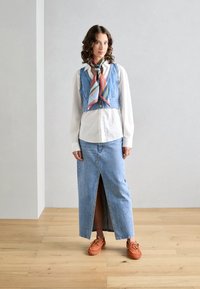 Gilet di denim con bottoni sopra una camicia bianca, abbinato a una lunga gonna di denim con uno spacco frontale. Accessoriato con una sciarpa multicolore e scarpe arancioni.