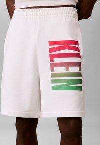 Short blanc avec taille élastique affichant un logo vertical "KLENN" en dégradé rouge-vert sur la cuisse gauche, porté par une personne à la peau foncée.