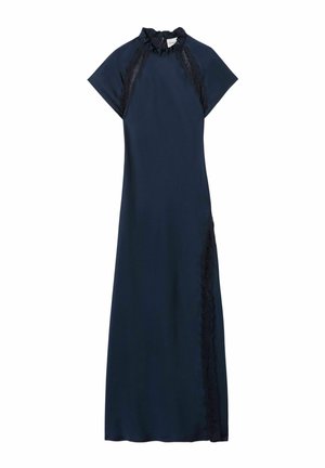 Giovane donna che indossa un abito lungo blu navy con dettagli in pizzo, tiene una piccola borsa bianca e indossa mocassini marroni e un braccialetto nero.