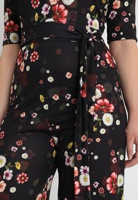Combinaison floreale avec un fond noir et une variété de fleurs colorées, ceinture en tissu à nouer et texture douce. Des manches mi-longues sont visibles.