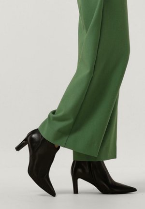 Personne portant un pantalon vert à jambes larges et des bottines noires à talons pointus, avançant sur un fond clair.