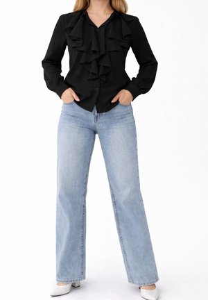 Mujer que lleva una blusa negra de manga larga con volantes, vaqueros de pierna ancha azul claro, tacones bajos blancos, de pie con las manos en los bolsillos sobre fondo blanco.