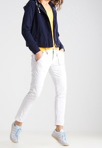 Marinblå kort jacka, gul V-ringad tröja, vita slim-fit byxor och ljusblå sneakers med vita detaljer och en texturerad sula.