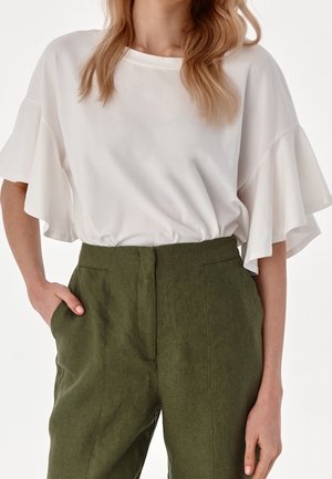 Woman wearing a white blouse with ruffled short sleeves tucked into high-waisted olive green pants with hands in pockets.