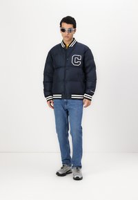 Námornícka prešívaná bunda s bielymi akcentmi a logom 'C', skombinovaná s modrými džínsami a sivými teniskami. Model má na sebe slnečné okuliare.