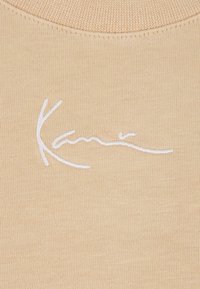 Beige neulotekangas, jossa on pyöreä pääntie, ja jonka pinnassa on valkoista brodeerattua tekstiä tyylitellyllä fontilla. Pehmeä pinta ja sileä viimeistely.