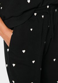 Schwarze Jogginghosen mit weißen Herzmustern, ausgestattet mit einem elastischen Bund und Seitentaschen. Weiches Baumwollmaterial mit lockerer Passform.