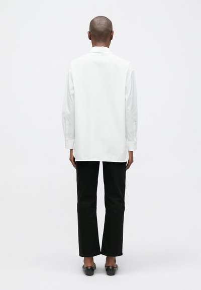 Biely dlhý rukáv košeľa so rovnými strihom, zadným yoke a gombíkmi na manžetách, v kombinácii s čiernymi nohavicami so rovnými nohavicami a plochými topánkami.