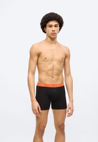 Μαύρα ανδρικά βαμβακερά μποξεράκια με πορτοκαλί ελαστική μέση που φέρει το λογότυπο "TOMMY HILFIGER", φορεμένα από μοντέλο με ουδέτερη έκφραση.