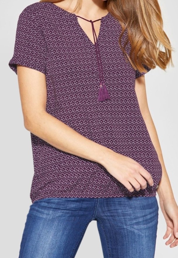 Femme portant un chemisier violet à manches courtes avec un motif, un col à lien avec pompons et un jean en denim bleu.