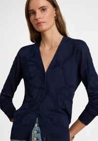 Tmavomodrý pletený kardigán s výstrihom do V, dlhými rukávmi a zapínaním na gombíky. Textúrovaný vzor pridáva detail tkanine.