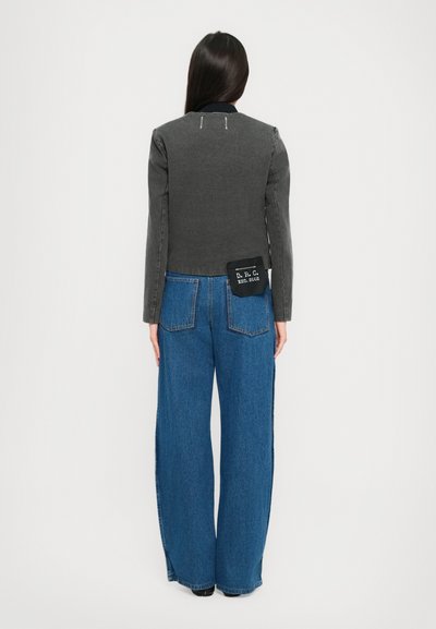 Vrouw die wegkijkt, gekleed in een donkergrijze jas en brede blauwe spijkerbroeken met een zwart label op de achterzak met "D.R.C EST. 2002".