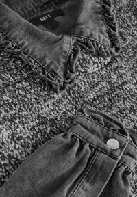 Grå teksturert strikket stoff med en frynset krage og sammenkrøllet denim-detalje. Har en sølvknap på siden. Merkemærket er synlig.
