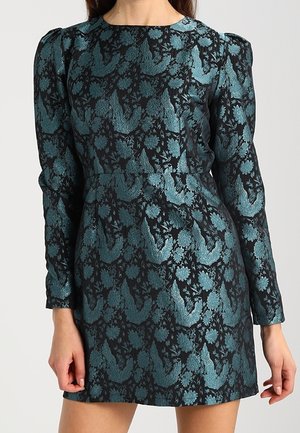 Vrouw draagt een aansluitende, korte jurk met lange mouwen, bedrukt met een jacquardpatroon van turquoise bloemen en vogels op zwarte stof.