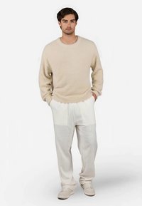 Suéter beige suave con escote redondo, combinado con pantalones de lino blancos que tienen acentos en panel. La modelo está de pie sobre un fondo simple.