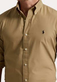 Koszula zapinana na guziki, wykonana z beżowej bawełny, z klasycznym kołnierzykiem, białymi guzikami oraz małym czarnym logo na piersi. Krótkie rękawy.