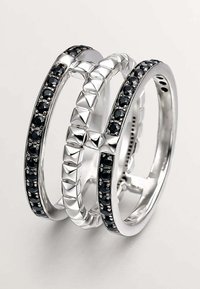 Tres anillos interconectados de plata con gemas negras. Un anillo tiene una superficie lisa, mientras que otro presenta acentos en forma de pirámide.