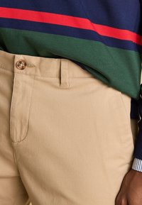 Pantalon beige avec bouton et passants pour ceinture, porté avec un pull vert marine comportant des rayures horizontales rouges et vertes, et un poignet visible d'une chemise rayée.
