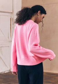 Suéter de punto rosa brillante con puños y dobladillo acanalados, con un ajuste relajado. Textura suave, mangas largas y cuello redondeado.