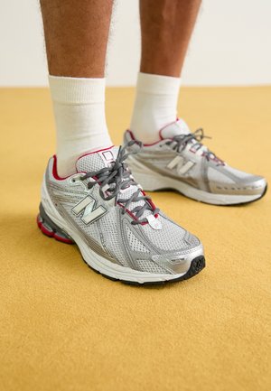 Stříbrné síťované sportovní boty s červenými akcenty, vybavené polstrovaným límcem a šedými tkaničkami, položené na žlutém texturovaném povrchu.