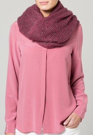 Femme portant une écharpe infinie en grosse maille bordeaux par-dessus un chemisier rose à manches longues, debout avec une main dans la poche.