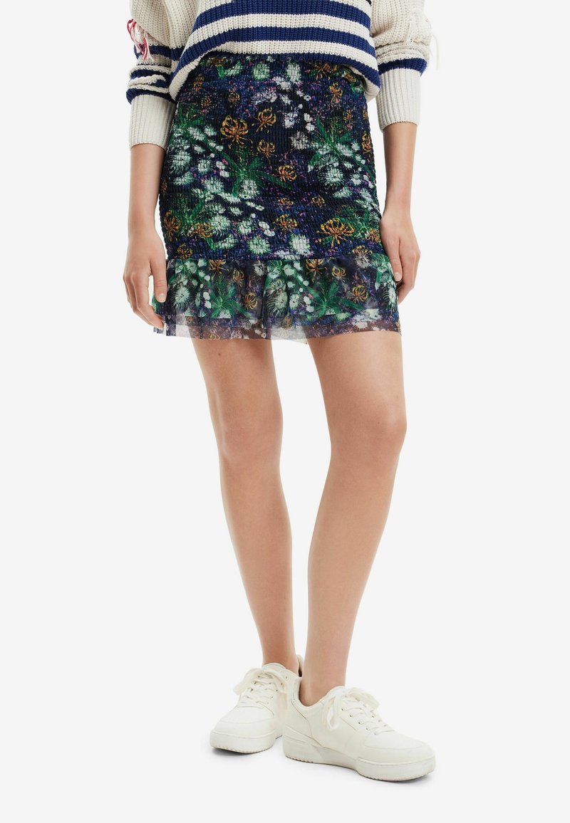 Pull tricoté à rayures bleues associé à une jupe courte à motif floral et des baskets blanches sur une personne debout.
