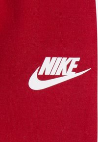 Κόκκινο ύφασμα με υφή στην επιφάνεια· διαθέτει ένα έντονο λευκό λογότυπο Nike με έντονο γραμματοσειρά και ένθετο σήμα swoosh.