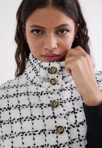 White and black textured jacket with a high collar, featuring gold accent buttons and a checkered pattern. Close-up view of the collar area.