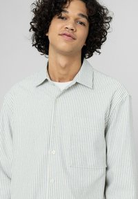 Chemise à boutons de couleur claire avec des rayures bleues verticales, en tissu à texture douce ; comprend une poche sur la poitrine gauche.