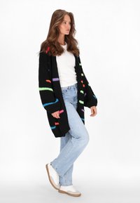 Melnā adīta jaka ar krāsainiem zigzaga rakstiem, brīvas formas, atvērta priekšpuse, apvienota ar gaiši ziliem džinsiem un baltām sporta kurpēm.