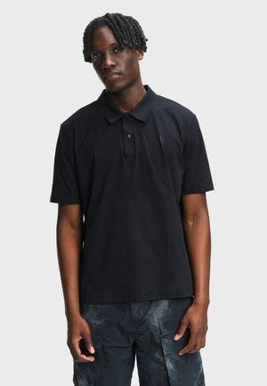 Homme aux dreadlocks courtes portant un polo noir à manches courtes et un pantalon cargo sombre à motifs, debout devant un fond clair uni.