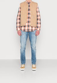 Gilet en toile beige porté sur une chemise à carreaux orange, bleu et blanc, associé à un jean bleu clair et des baskets marron clair avec des semelles blanches.