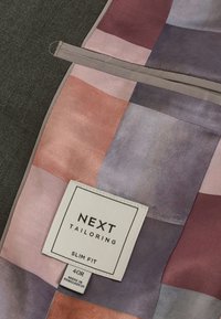 Tissu de vêtement sur mesure en blocs rose, violet et gris avec une étiquette blanche indiquant "NEXT Tailoring Slim Fit 40R Made in Bangladesh."
