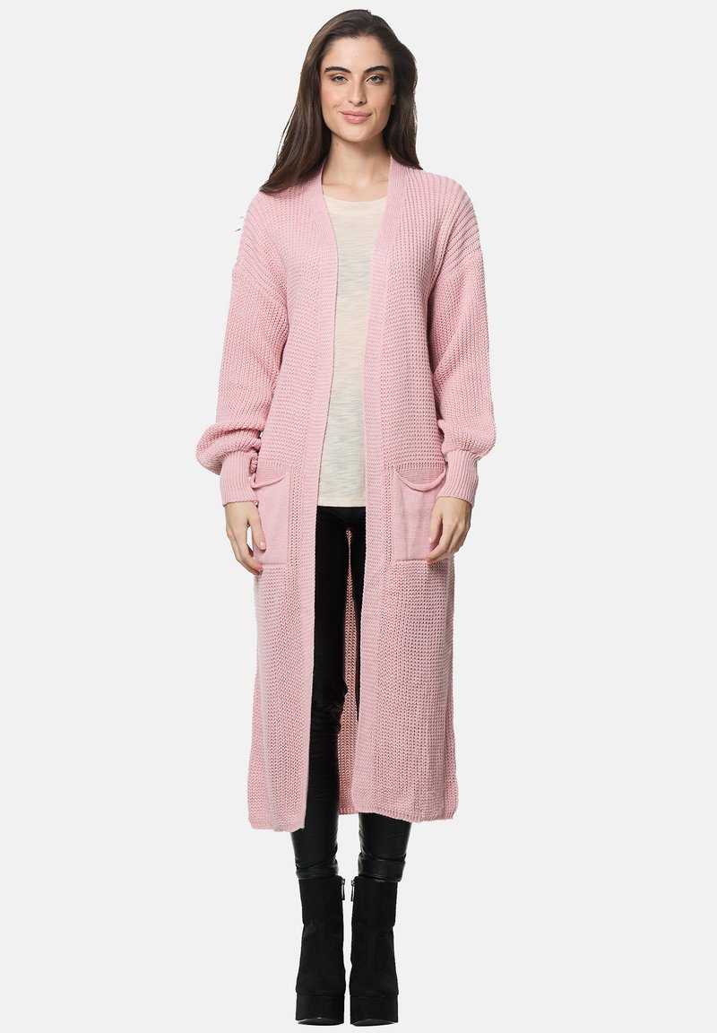 Lång rosa stickad cardigan med öppen front, ribbad textur, sidfickor och åtsittande ärmar, bärs över en ljus topp och svarta byxor.