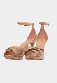 Μπεζ σανδάλια ανοικτού πέλματος με ψηλό τακούνι, μπροστινές λουρίδες δεμέ νες σε κόμπο και κλεισίματα με αγκράφα στον αστράγαλο σε λευκό φόντο.