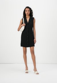 Black sleeveless dress with a collar, front buttons, and side pockets. White handbag with a rounded shape, paired with beige open-toe heels.