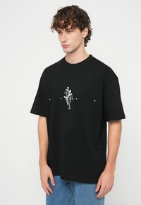 Νέος άνδρας με σγουρά μαλλιά που φοράει υπερμεγέθη μαύρη μπλούζα με γράφημα και κείμενο, συνδυασμένη με μπλε τζιν, στέκεται μπροστά σε απλό φόντο.