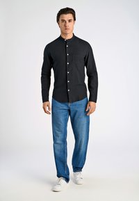 Camicia nera con colletto, tasca singola sul petto, abbinata a jeans denim blu e sneakers bianche. Design semplice e casual.