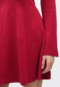 Piros ruha hosszú ujjal, bővülő szoknyával és texturált anyagból. Lekerekített aljú, bordázott szegéllyel. Nincsenek látható minták vagy díszítő elemek.