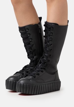 Bottes plateformes noires montantes jusqu'au genou avec semelles épaisses côtelées, lacets sur le devant et fermetures éclair sur le côté, portées sur des jambes nues sur un fond blanc.