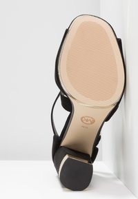 Chaussure à talon haut noire avec une finition en suède texturé, dos ouvert et un talon fin accentué d'or. La semelle présente un motif en relief.