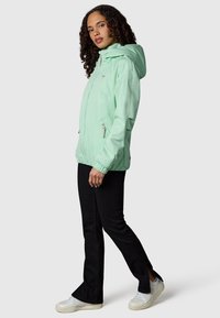 Vrouw draagt lichtgroen gewatteerd jack met capuchon, zwarte broek met gespleten zoom en witte sneakers, staand in een driekwarts profiel.