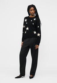 Donna con trecce lunghe che indossa un maglione nero con stelle bianche, pantaloni neri a gamba larga e scarpe nere con punta, in piedi contro uno sfondo semplice.