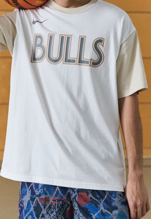 Personne portant un maillot de basketball des Bulls blanc et beige tenant un ballon de basketball orange sur son épaule, short bleu à motifs visible.