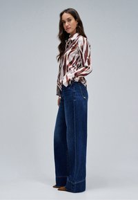 Frau mit langen Haaren, die eine braun-weiß gemusterte Bluse, weite dunkelblaue Jeans und braune spitze Schuhe vor grauem Hintergrund trägt.