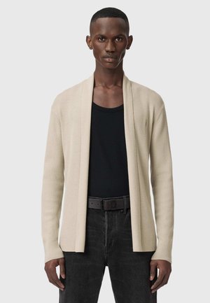 Homme portant un cardigan beige ouvert sur une chemise noire, associé à un jean noir et une ceinture noire, debout devant un fond gris.