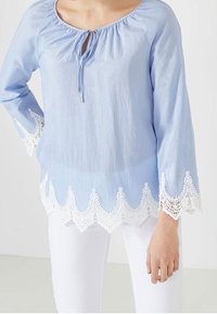 Ljusblå blus med långa ärmar, rund halsringning, knytdetalj fram, och vit spets- nederdel med vågiga kanter. Slät tygtextur.